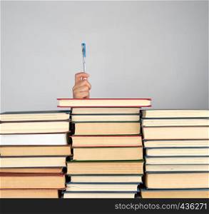 stack of different books on a gray background, hand sticks out from behind a pile and holding blue pen