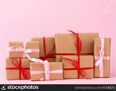 stack of gifts in boxes wrapped in brown kraft paper and tied with silk ribbon on a pink background. Festive concept, copy space