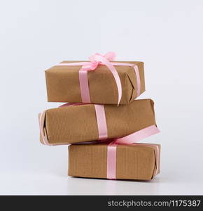 stack of gifts in boxes wrapped in brown kraft paper and tied with pink silk ribbon on a white background. Festive concept, copy space