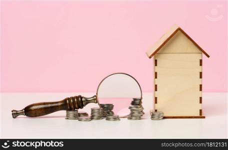 stack of metallic money and magnifier and wooden house on a pink background. Real estate rental, purchase and sale concept. Realtor services, building repair and maintenance