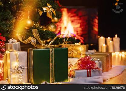 Stack of packed gift boxes under Christmas tree against burning fireplace. Lots of Christmas gifts under the tree. Candles on wooden floor. Focus on green box!