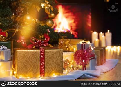 Stack of packed gift boxes under Christmas tree against burning fireplace. Lots of Christmas gifts under the tree. Candles on wooden floor. Focus on golden box!