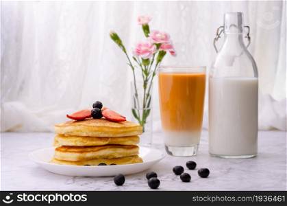 Stack of pancakes with fresh strawberries and blueberries on a white plate, a bottle of milk, a glass of milk tea and a vase of pink flowers on marble table. Healthy morning breakfast.