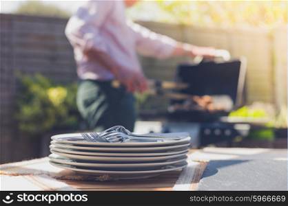 Stack of plates on a table outside in a garden with a man attending to a barbecue in the background