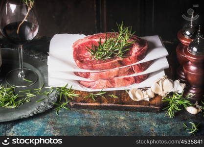 Stack of raw steaks with rosemary ,spices and grass of red wine on kitchen table at dark wooden background, side view