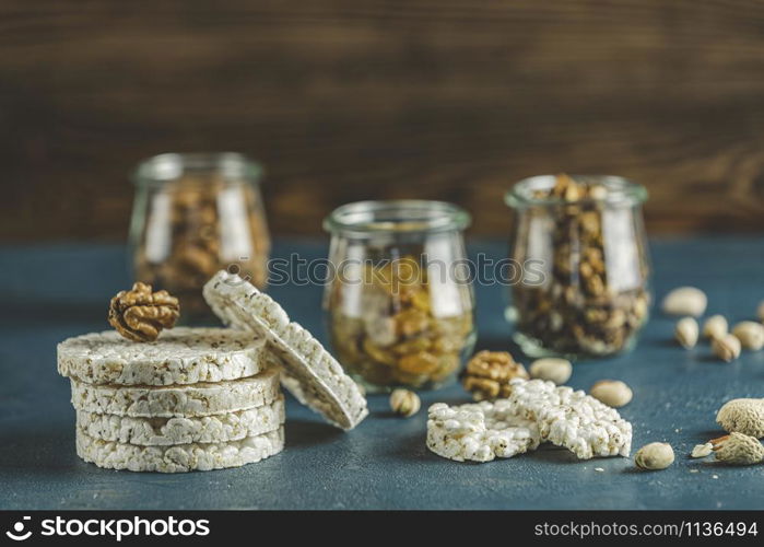 Stack of rice cakes. American puffed rice cakes. Healthy snacks with almonds, raisins, peanuts, pistachios in glass jars on classic blue concrete surface. Close up.