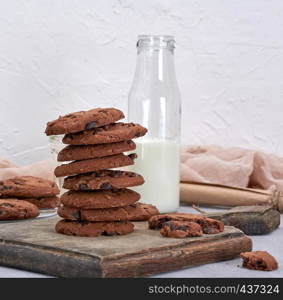 stack of round chocolate chip cookies on a brown wooden board, behind a bottle of milk