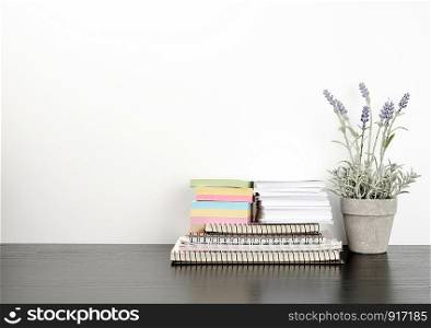stack of spiral notebooks and colored stickers, next to a ceramic pot with a flower on a black table, white wall, Scandinavian-style minimalism