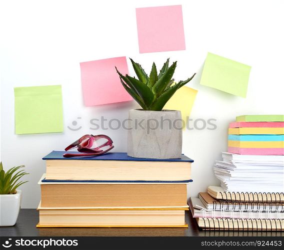 stack of spiral notebooks and colored stickers, next to a ceramic pot with a flower on a black table, white wall