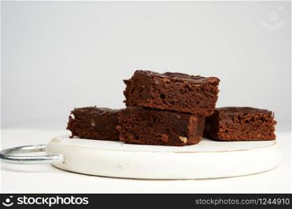 stack of square baked slices of brownie chocolate cake with walnuts on a round white wooden board, close up
