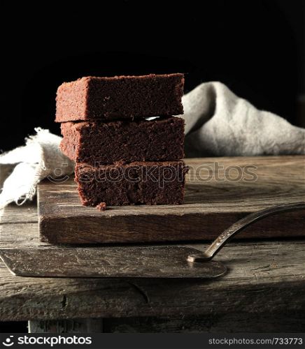 stack of square pieces of baked brown brownie pie on a wooden board, black background