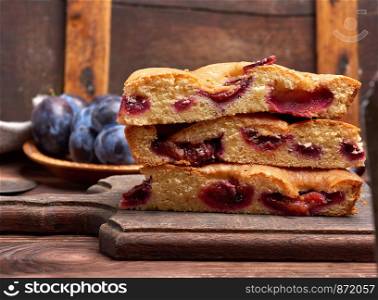 stack of triangular slices of sponge cake with blue plums on a vintage brown board, wooden table