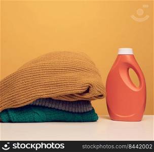 stack of washed folded clothes and plastic orange large bottle with liquid detergent stand on a white table, yellow background. Routine homework