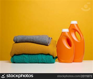 stack of washed folded clothes and plastic orange large bottles with liquid detergent stand on a white table, yellow background. Routine homework