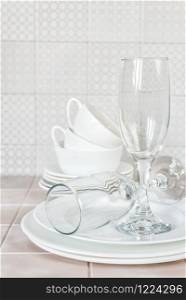 Stack of white porcelain plates, tea cups and wine glasses on the kitchen table against the background of a apron made of ceramic tile