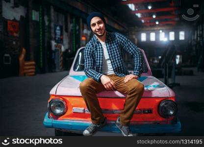 Stalker, male traveler pose against retro car, factory interior on background. Danger zone, mysterious adventure in deserted place