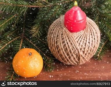 standing candle in a homemade candlestick, mandarin and fir branches on a wooden surface. Falling snow