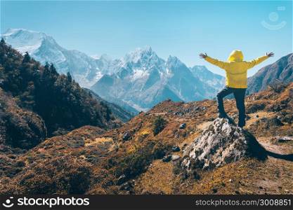 Standing man with raised up arms on the stone and looking on snow covered mountains. Landscape with traveler, high rocks with snowy peaks, grass, trees in autumn in Nepal. Lifestyle, travel. Trekking