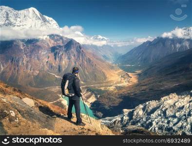 Standing sporty man with backpack on the mountain peak and looking on beautiful mountain valley at sunset. Landscape with man, rocks with snowy peaks, glacier, clouds in Nepal. Hiking, travel.Trekking. Standing sporty man with backpack on the mountain peak