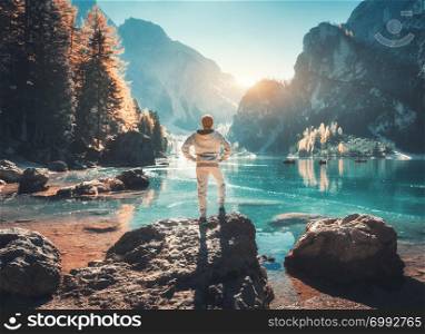 Standing young man on the stone on the coast of Braies lake at sunset. Autumn in Dolomites, Italy. Landscape with happy man, mountains, water with reflection, fall trees and blue sky in the evening