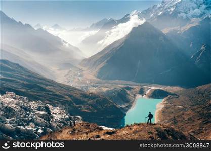 Standing young woman on the mountain peak and looking on beautiful mountain valley with lake, river and hills at sunset. Landscape with girl, rocks in clouds, sky, glacier in Nepal. Travel. Trekking