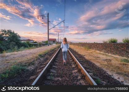 Standing young woman on the railroad at sunset in summer. Slim girl on the railway station. Rural industrial landscape with railroad, sky with colorful clouds. Railway platform. Transportation. Travel. Standing young woman on the railroad at sunset in summer