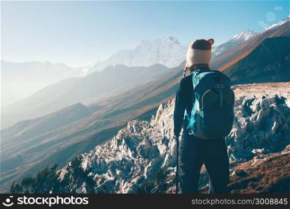 Standing young woman with backpack on the mountain peak and looking on beautiful mountain valley at sunset. Landscape with girl, rocks with snowy peaks, hills, blue sky in Nepal.Hiking, travel.Vintage