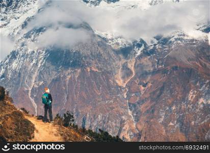 Standing young woman with backpack on the path on the hill and looking on beautiful high mountains at sunset. Landscape with girl, rocks with snowy peaks, low clouds in Nepal. Hiking, travel. Trekking. Young woman with backpack on the mountain peak