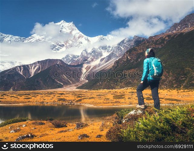 Standing young woman with backpack on the stone and looking on mountains in clouds at sunset. Landscape with girl, high rocks with snowy peaks, yellow grass, blue sky in Nepal. Travel. Trekking