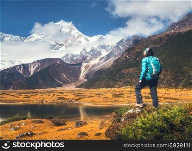 Standing young woman with backpack on the stone and looking on mountains in clouds at sunset. Landscape with girl, high rocks with snowy peaks, yellow grass, blue sky in Nepal. Travel. Trekking