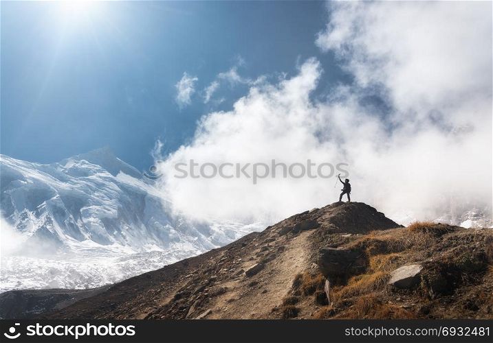 Standing young woman with raised up arms on the mountain peak against blue sky with low clouds in bright day. Landscape with happy girl on the hill, snowy mountains in Nepal. Travel in Himalayas. Young woman with raised up arms on the mountain peak
