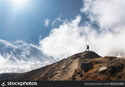 Standing young woman with raised up arms on the mountain peak against blue sky with low clouds in bright day. Landscape with happy girl on the hill, snowy mountains in Nepal. Travel in Himalayas. Young woman with raised up arms on the mountain peak