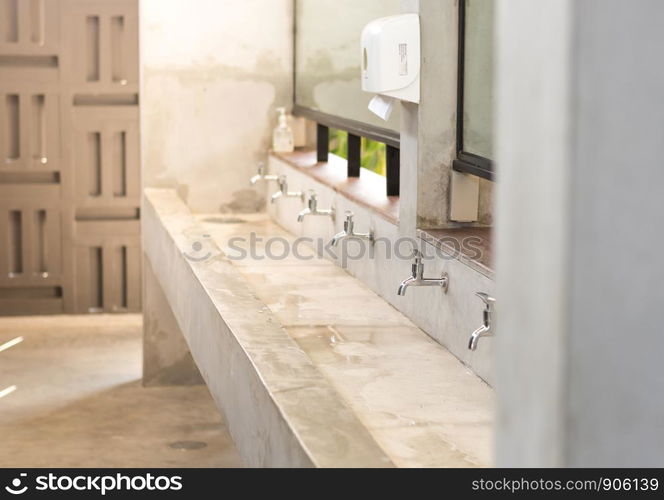 Stanless steel taps above concrete sink in shared toilet with yellow sunlight for background.