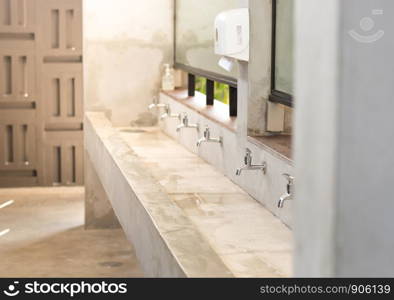 Stanless steel taps above concrete sink in shared toilet with yellow sunlight for background.