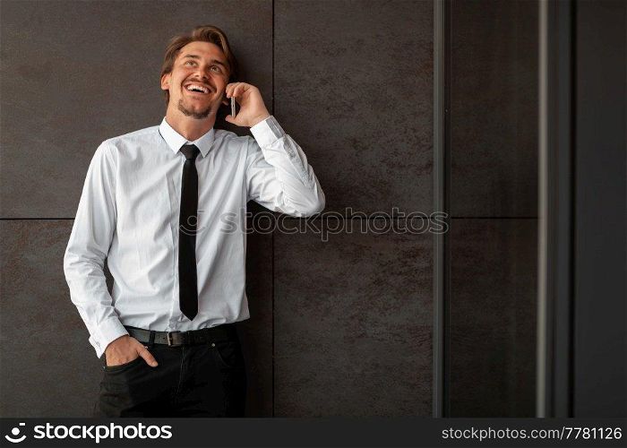 Startup businessman in a white shirt with a black tie using smartphone while standing in front of gray wall during a break from work outside. High-quality photo. Startup businessman in a white shirt with a black tie using smartphone while standing in front of gray wall during break from work outside