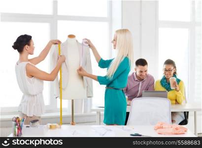 startup, education, fashion and office concept - smiling fashion designers measuring and decorating jacket on mannequin