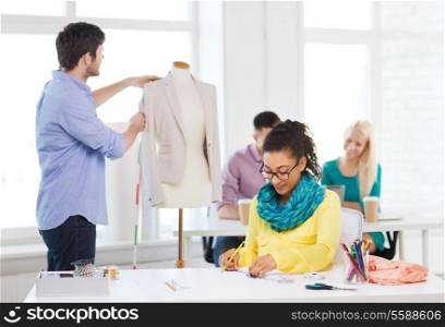 startup, education, fashion and office concept - smiling female drawing sketches and male measuring jacket on mannequin in office