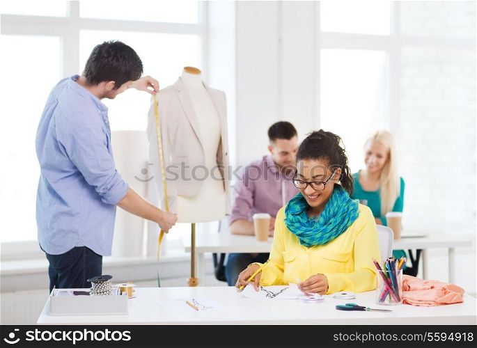 startup, education, fashion and office concept - smiling female drawing sketches and male measuring jacket on mannequin in office