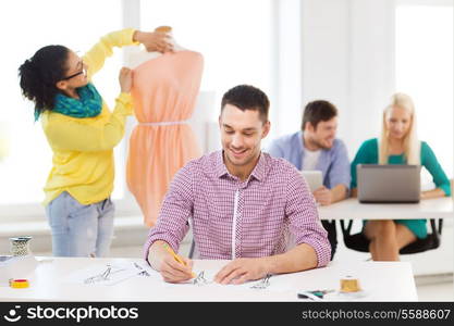 startup, education, fashion and office concept - smiling male drawing sketches and female adjusting dress on mannequin in office