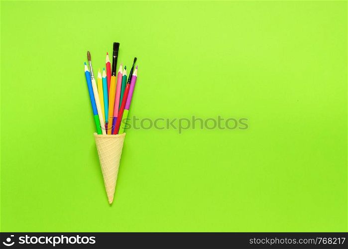 Stationery colored Pencils paintbrush in waffle ice cream cone. Still life on green background. Copy space Flat lay Top view Concept Education . tationery colored Pencils paintbrush in waffle ice cream cone