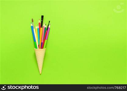 Stationery colored Pencils paintbrush in waffle ice cream cone. Still life on green background. Copy space Flat lay Top view Concept Education . tationery colored Pencils paintbrush in waffle ice cream cone