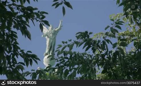 Statue of Victoria against blue sky, view through the green tree branches. The Roman goddess of Victory