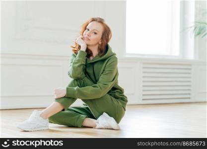 Staying in good shape. Pleasant looking red haired female gymnast with wavy hair, sits on floor in fitness hall, has flexible body, rests after doing yoga exercises, looks confidently at camera