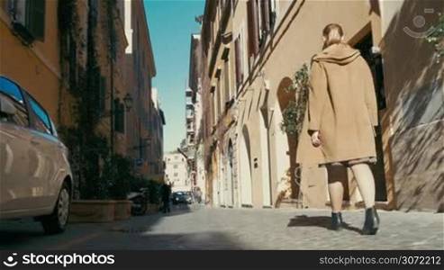 Steadicam and low angle shot of a woman in a coat walking along the old narrow street in Europe. She looking upon the old architecture