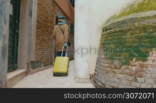 Steadicam back shot of a woman in coat rolling trolley bag along the narrow passage between old worn buildings in Italy. Traveling in Europe