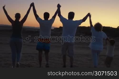 Steadicam back shot of big family of parents, little son and grandparents walking on the beach at sunset. The holding hands and then raising them. Happy vacation together