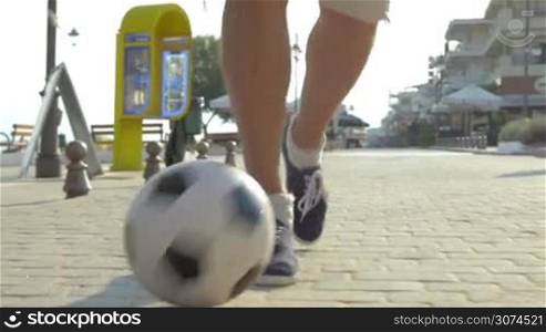 Steadicam close-up shot of man dribbling a soccer ball on the paved sidewalk in resort town, street cafes and hotels in background