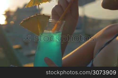 Steadicam close-up shot of young blond woman in summer hat having cocktail with ice while relaxing on the beach at sunset