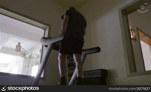 Steadicam low angle shot of a mature man exercising on treadmill working on low speed. Gym in the hotel with view on the terrace