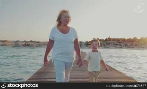 Steadicam shot of a grandmother walking with little grandchild along the wooden pier on resort. They holding hands, woman talking to the boy and they looking at sea